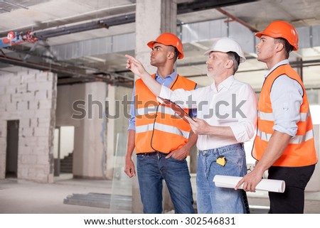 Experienced old architect is explaining to construction team the concepts of project. He is pointing his finger sideways seriously. The workers are looking there with interest. Copy space in left side