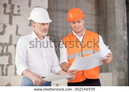 Senior architect and young builder are discussing the new project. They are holding blueprints and looking at it with interest. The foreman is smiling