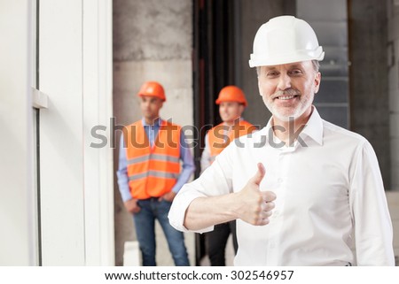 Cheerful old architect is giving his thumbs up. His project was done perfectly. The man is smiling and looking at the camera happily. Two builders are standing behind him and relaxing