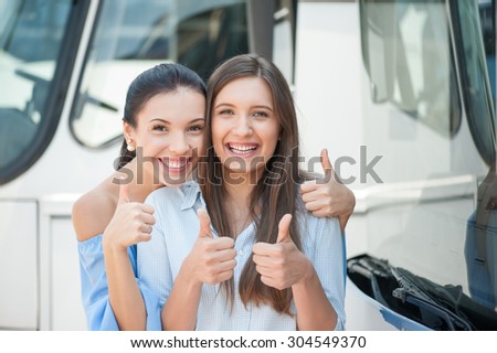 Cheerful women are standing near a bus. They are giving thumbs up and smiling. The friends are embracing. They are waiting for bus departure