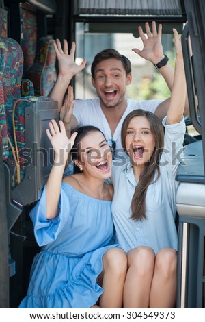 Cheerful friends are sitting on doorsteps of a bus. They are raising their hands up and smiling. The women are embracing. The man is sitting behind them