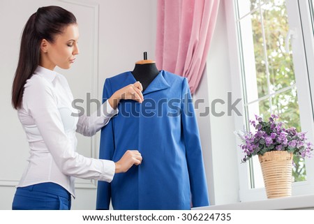 Pretty clothes designer is creating new clothing. She is adjusting it on mannequin with inspirations. The lady is looking at a jacket with concentration