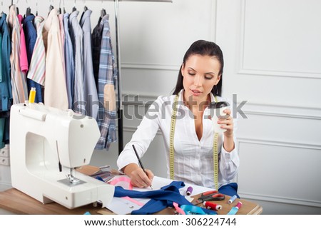 Cheerful clothes designer is working on a new collection. The woman is drawing sketches of fashionable clothes. She is looking at a blueprint with aspirations and drinking coffee
