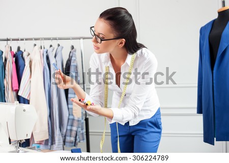 Pretty clothes designer is holding a set of threads. She is chooses on of them for sewing. The woman is looking at blue thread with aspirations. She has eyeglasses and tape-measure