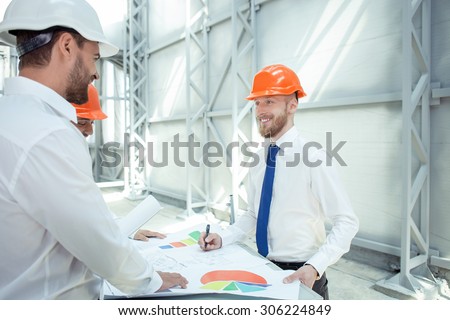 Attractive engineers are planning the new building. They are working with sketches and standing near the table. The men are smiling and looking at each other with trust