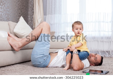 Handsome young man is lying on flooring and smiling. He is splaying with his child with pleasure. The boy is sitting on him with joy. There is a tablet on the carpet