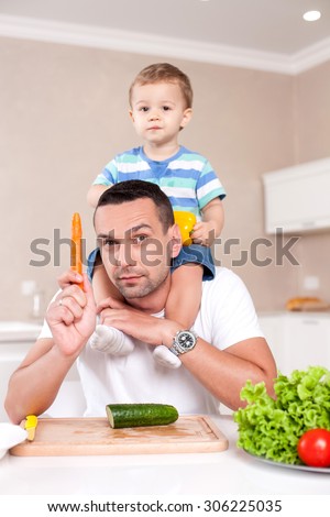 Handsome man is teaching his son to eat healthy food. He is sitting at the table and holding toddler on his shoulders. The man is showing carrot and smiling. The boy is holding a pepper with interest