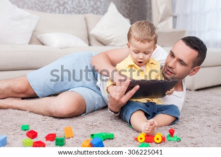 Cheerful young man is lying on flooring near his little son. The boy is sitting on carpet and holding a tablet. The child is looking at it with amazement. His father is smiling