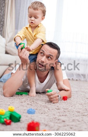 Handsome young father is playing with his son. He is holding the kid on his shoulders and giving his toys. The man is lying and smiling. A boy is taking playthings with joy