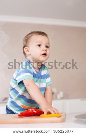Cute small boy is standing near a table in the kitchen. He is touching chopped vegetable with appetite. The toddler is looking forward with interest