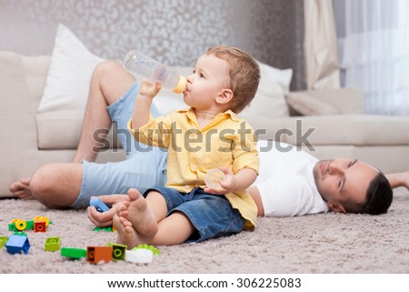 Handsome father is lying on flooring near his sun. He is looking at kid with love and gently smiling. The boy is sitting and drinking water from a bottle with concentration