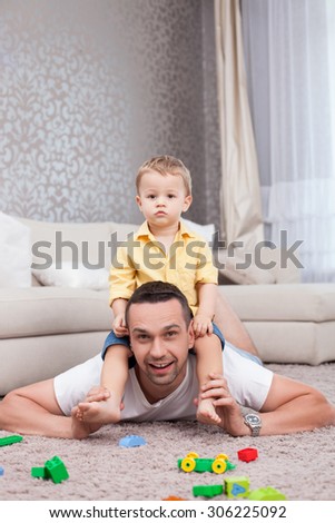 Cheerful man is playing with his son. He is lying on flooring and holding a kid on his shoulders. The man is looking at the camera and smiling. A boy is sitting with joy