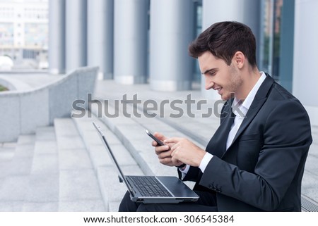 Cheerful man in suit is sitting on steps. He is holding a mobile phone and messaging to his friend. The man is smiling. There is a notebook on his knees