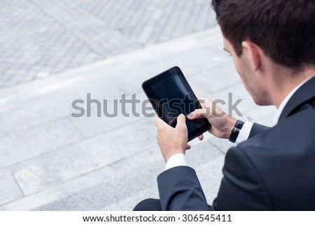 Successful male worker is holding a tablet in his hands. He is touching it and looking at it with joy. The man is sitting on steps. Copy space in left side