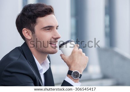 Cheerful man in suit is holding a cup and drinking coffee. He is sitting on steps near his office. The man is smiling and looking forward with enjoyment. Copy space in right side