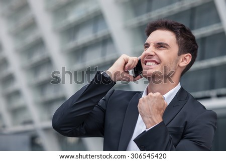 Handsome businessman is standing and talking on the phone with his client. The deal was done successfully. The worker is smiling and raising his fist up happily