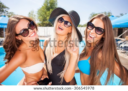 Attractive women are posing and smiling. They are standing near a swimming pool in swimwear and sunglasses. The friends are looking at the camera with joy