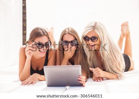 Beautiful women are lying in swimsuits. They are looking at the laptop and laughing. The brunette is holding a technology and covering her moth with shock