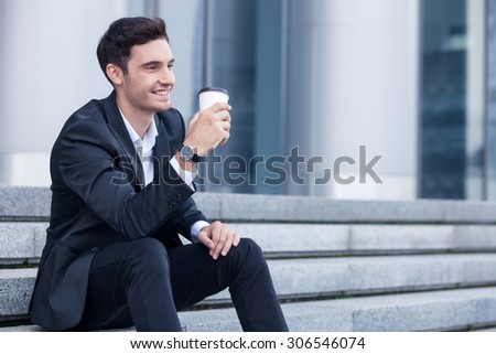 Attractive young businessman is sitting on steps outdoors. He is drinking coffee and smiling. The guy is looking aside with joy. Copy space in right side