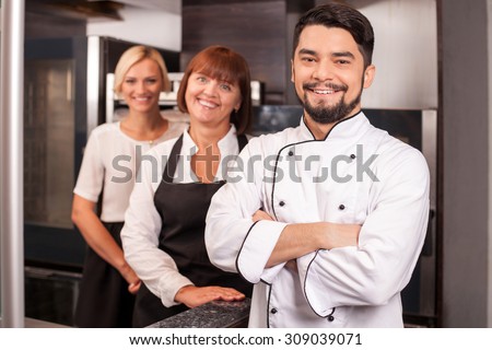 Attractive young male baker and his assistants are standing in kitchen. They are smiling happily. The cooking team is looking at the camera happily