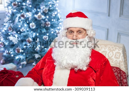 Happy New Year. Cheerful Santa Claus is sitting in his chair near a fir-tree and a red sack. He is looking forward and smiling with happiness