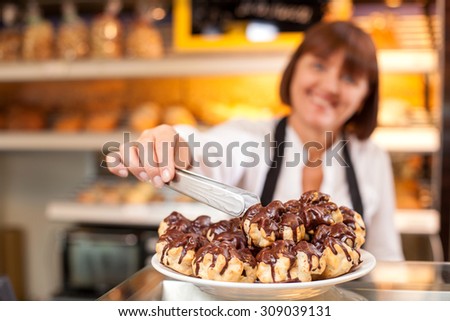 Cheerful saleswoman is standing at the counter in bakehouse. She is taking sweet cookie from the plate. The woman is smiling. Focus on pastry