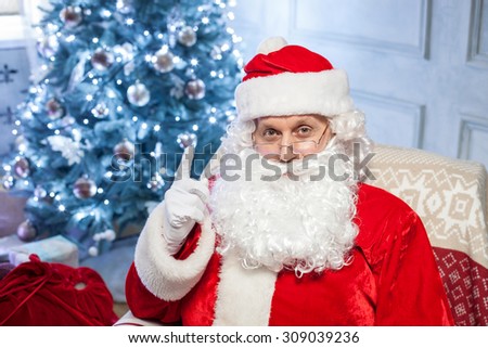 Gorgeous Father Christmas is sitting in chair and pointing his finger up. He is teaching children to be good. The man is looking at the camera seriously. There is Christmas tree on background