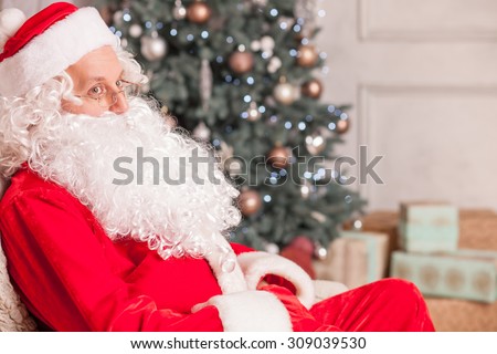 Gorgeous Santa Claus is sitting on his chair and resting. He is looking side with seriousness. There are many boxes of presents and a holiday fir-tree on the background