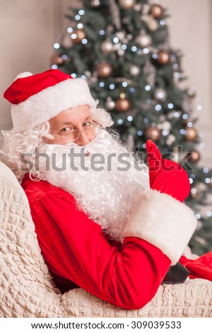 Old Father Christmas is giving thumb up and smiling. He is sitting in his chair near a Christmas tree. The man is looking at the camera with happiness