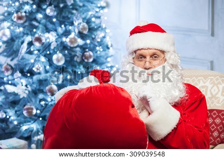 Bearded Santa Claus is preparing to give a gift. He is sitting in a chair near a holiday fir-tree and holding a red bag. The man is raising his finger to lips and asking for silence mysteriously