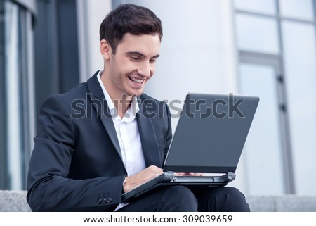 Handsome businessman is sitting on steps near his office. He is holding a notebook on his knees and looking at the screen with joy. The guy is smiling