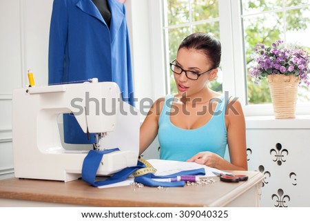 Beautiful fashion designer is looking at the sketches of clothing with concentration. She is sitting at the table near a sewing machine and smiling