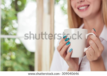 Close up of beautiful blonde girl is drinking coffee near a window. She is holding a cup and smiling. Copy space in left side