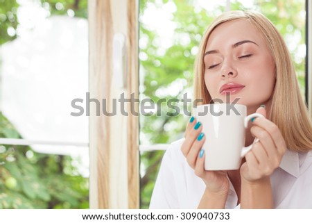 Pretty blond girl is drinking coffee near a window. She closed her eyes with pleasure. The lady is relaxing and smiling. Copy space in left side