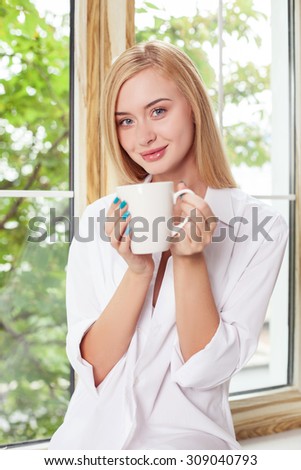 Beautiful girl is sitting on a windowsill. She is drinking a cup of coffee. The lady is looking forward and smiling. She is wearing a white male shirt