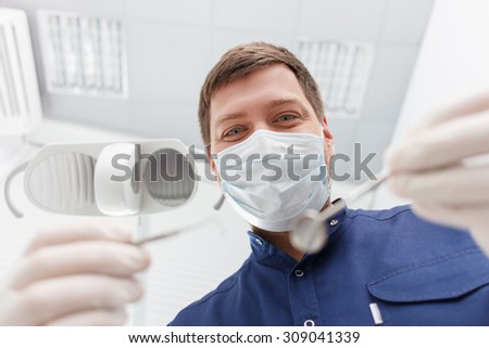 Experienced dental doctor is standing and showing the medical tools to the camera. He is looking forward happily. The man is wearing a mask and gloves