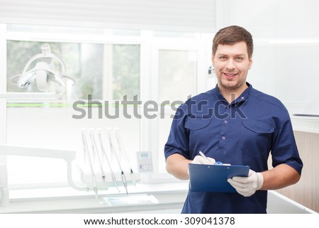 Attractive young dentist is standing and listening to the complaints of his patient. He is writing down the data and smiling. Copy space in left side