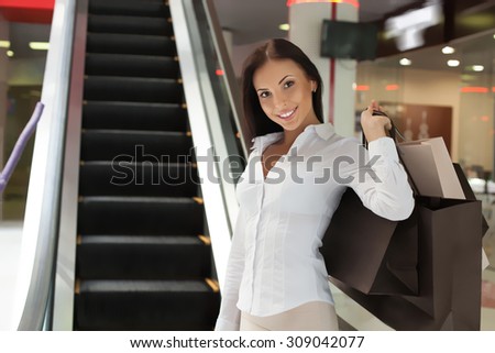 Cheerful girl is standing near excavator in shopping center. She is holding many packages of bought clothing and raising it behind her back. The lady is smiling. Copy space in left side