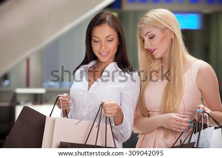 Beautiful girls are standing in boutique and holding many packages. One lady is opening her packet and showing new clothing to her friend proudly. They are smiling