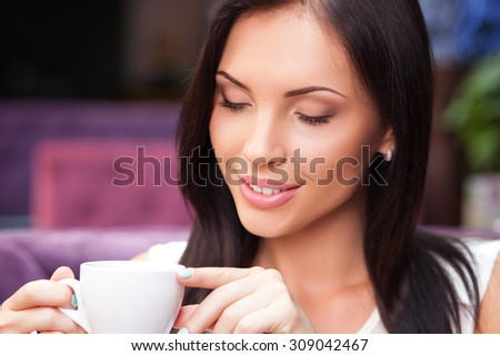 Attractive girl is drinking coffee and smiling. She is sitting and relaxing in cafeteria. Her eyes are closed with pleasure