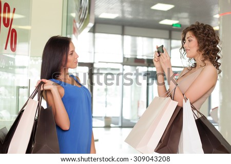 Beautiful women are making fun in shop. They are standing with joy and holding many packets of bought clothing. One woman is photographing her friend on the mobile phone and smiling