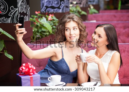 Attractive women are making selfie in cafe. They are sitting at the table and drinking coffee. The friends are looking at the mobile phone and smiling. There is a box of gift on the table