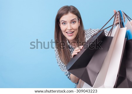 Cheerful styled woman is holding many packages of bought clothing and smiling. She is hiding behind it and peeking through it with joy. Isolated on blue background and copy space in left side