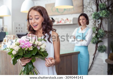 Attractive woman is holding beautiful flowers in store. She is looking at it with amazement and laughing. The florist is looking at her customer happily and smiling
