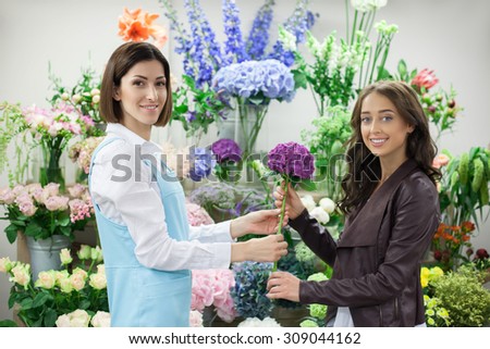 Beautiful saleswoman is giving a flower to her female client. The women are looking at the camera with joy and smiling. They are standing near a lot of bouquets in flower shop