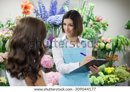 Professional florist is taking with her client and writing down her order. The woman is looking at the girl positively and smiling