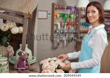 Cheerful florist is making a bouquet in her workshop. She is touching roses happily and smiling. The woman is looking at the camera with inspiration