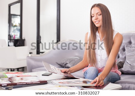 Attractive girl is waiting for hairdresser in beauty shop. She is sitting on sofa and reading magazines with interest. The lady is smiling. Copy space in left side