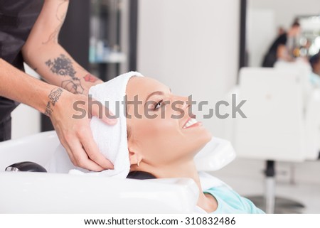Beautiful woman has her hair washed in salon. She is leaning her head on a sink and smiling. The male hairdresser is drying female hair with a towel