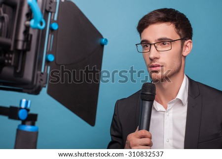 Attractive reporter is holding a microphone and telling news. The man is looking at the camera seriously. He is wearing suit and eyeglasses. Isolated on blue background
